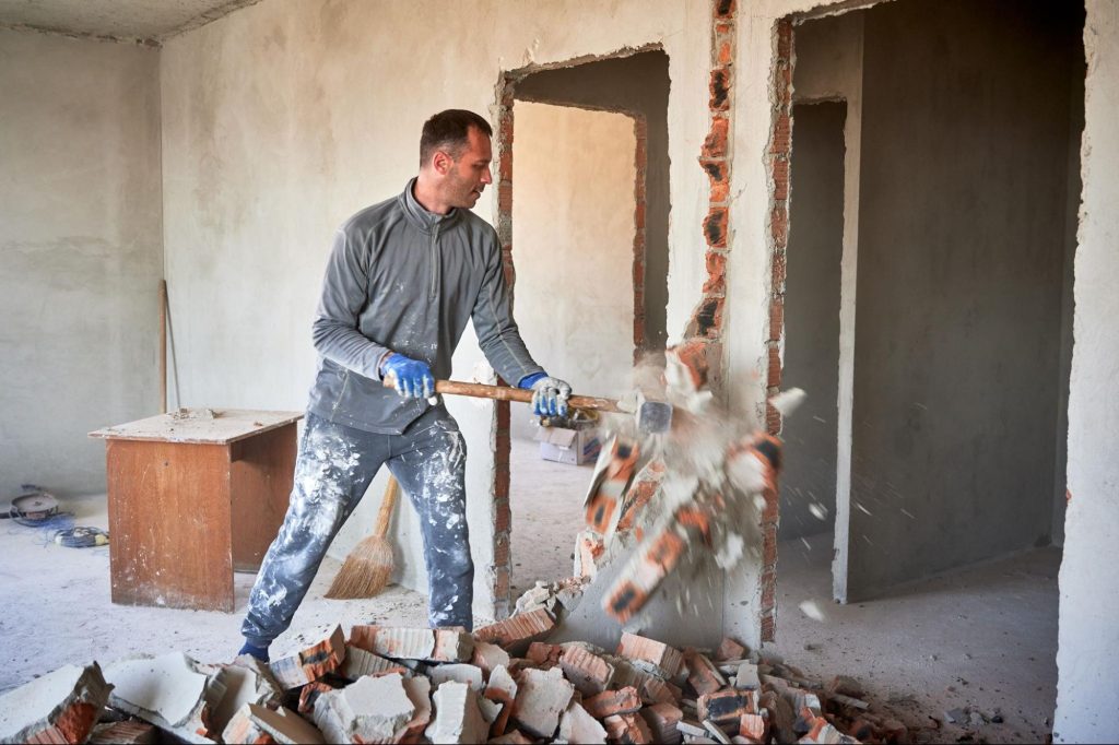 Man breekt met een voorhamer een bakstenen muur af tijdens een renovatie binnenshuis.
