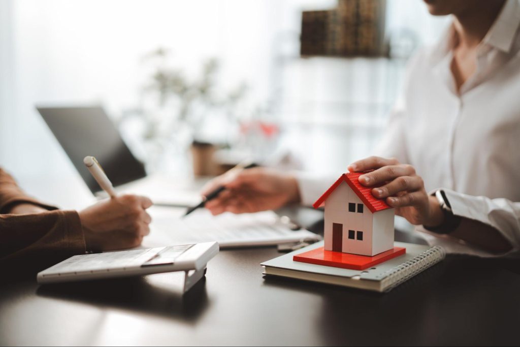 Adviseur en klant bespreken aan tafel de financiering van een woning met modelhuisje en rekenmachine.
